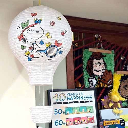A white paper hot air balloon lantern featuring Snoopy holding a tennis racket surrounded by colorful butterflies. Below it sits a framed poster reading "40 Years of Happiness Peanuts", along with a framed Charlie Brown latch-hook craft and a plush Woodstock in the background.