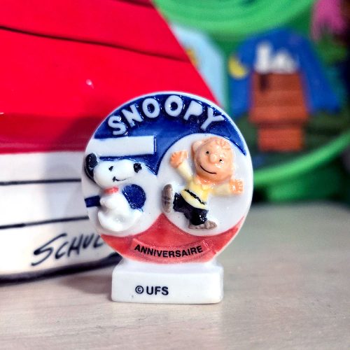 A king cake trinket featuring a circular 50th Anniversary badge with Snoopy and Charlie Brown set against a red, white, and blue backdrop. The top of the badge reads "SNOOPY", the red bottom banner reads "ANNIVERSAIRE", and the white base is printed with "© UFS". Snoopy's doghouse is softly blurred in the background.