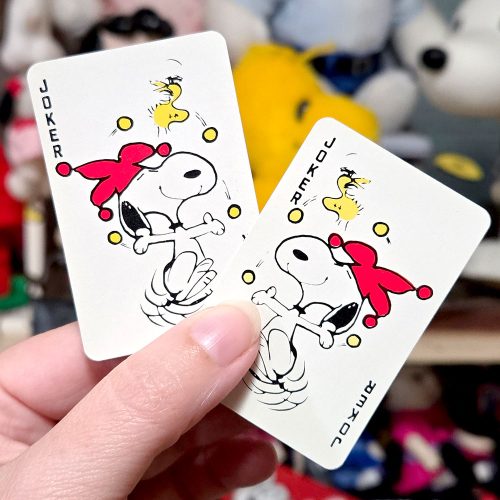 Fingers holding two Peanuts playing cards featuring Snoopy and Woodstock dressed as jokers, with a blurred collection of plush toys in the background.
