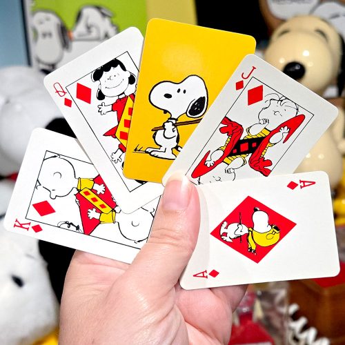 A hand holding four Peanuts playing cards featuring diamonds, including an Ace with Snoopy inside the diamond pip, a King with Charlie Brown, a Jack with Linus, and an overlapping card showing Queen Lucy.