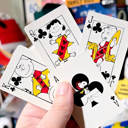 A hand holding four Peanuts playing cards featuring clubs, including an Ace with Snoopy inside the club pip, a King with Charlie Brown, a Jack with Linus, and an overlapping Queen with Lucy.