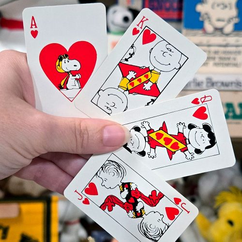 A hand holding four Peanuts playing cards featuring hearts, including an Ace with Snoopy inside the heart pip, a King with Charlie Brown, a Jack Linus, and Queen Lucy.
