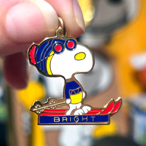 A metal enamel charm of Snoopy wearing a blue ski cap with red goggles and a yellow shirt, skiing on red skis across a blue base that reads "BRIGHT".