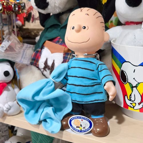 Linus 40th Anniversary Doll from Applause wearing his signature blue striped shirt and black shorts, holding his blue security blanket while seated next to a round commemorative button badge.