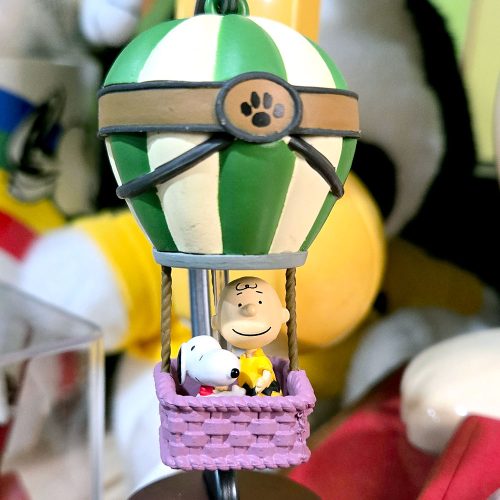 A miniature figurine of a green-and-white striped hot air balloon with a paw print detail, carrying Charlie Brown and Snoopy in a purple wicker basket.
