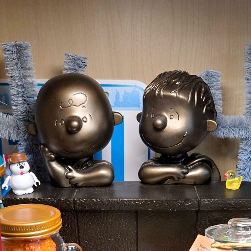 A metallic bronze-toned figurine of Charlie Brown and Linus leaning with their arms crossed, set against a background featuring blue tinsel and a small Olaf figure on the left.