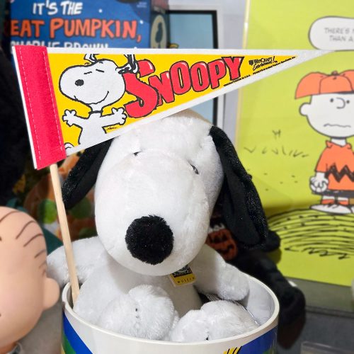 A plush Snoopy doll sits inside a blue and white container while holding a red felt pennant featuring an illustration of Snoopy and the bold yellow text "SNOOPY," with a Charlie Brown "It's the Great Pumpkin" cover visible in the background.