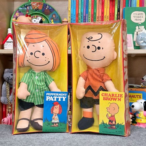 Two boxed Peppermint Patty and Charlie Brown rag dolls from Ideal stand side-by-side, featuring Peppermint Patty in her green striped shirt and shorts and Charlie Brown in his classic zigzag shirt, set against a bookshelf background.