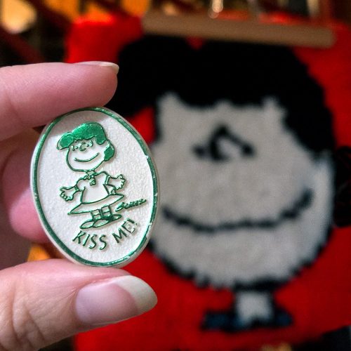 A hand holds an oval "Lucy 'Kiss Me'" emblem clip featuring an illustration of Lucy van Pelt with green outlines and text, against a blurry background showing a Lucy latch hook rug.
