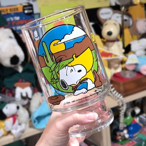 A hand holds up a clear glass pedestal vase featuring a colorful illustration of Cowboy Snoopy resting against a saddle in a desert landscape with a cactus and yellow sky.