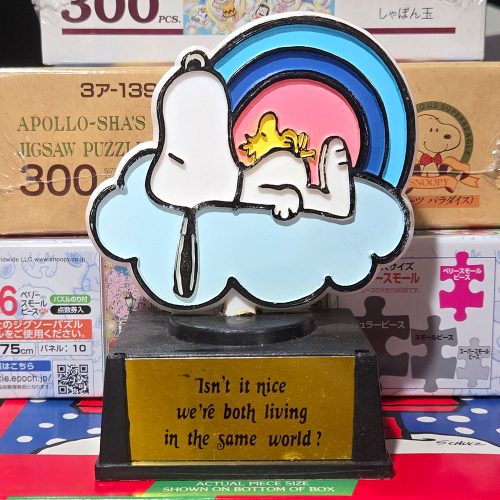 A Snoopy and Woodstock plaque featuring Snoopy lying on a blue-framed rainbow arch with Woodstock, set on a wooden base with an inscription that reads, "Isn't it nice we're both living in the same world?"