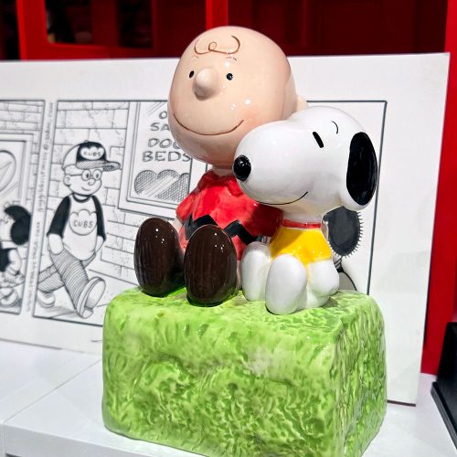 Vintage Peanuts figurine featuring Charlie Brown and Snoopy sitting side-by-side on a green, textured grassy bank base, set against a background showing comic strip panels.