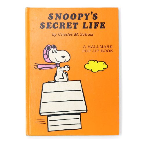 Cover of Snoopy's Secret Life Pop-Up Book by Charles M. Schulz against an orange background, featuring Snoopy dressed as the World War I flying ace with a pink scarf and aviator cap, sitting on top of his doghouse next to a yellow cloud, with the title text "Snoopy's Secret Life: A Hallmark Pop-Up Book."