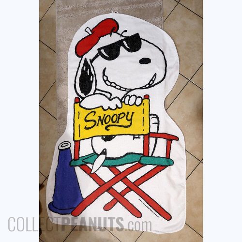 A vintage white beach towel featuring Snoopy wearing sunglasses and a red beret, sitting on a red-and-turquoise director's chair with a yellow back that reads "SNOOPY." Woodstock peeks out from behind the chair, a blue megaphone stands on the left, and a paintbrush is held in Snoopy's paw.