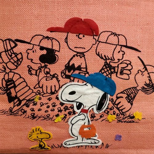 Embroidery or fabric panel featuring Snoopy wearing a blue baseball cap holding a glove, with Charlie Brown, Lucy, and other Peanuts characters gathered behind him on an orange-pink background.