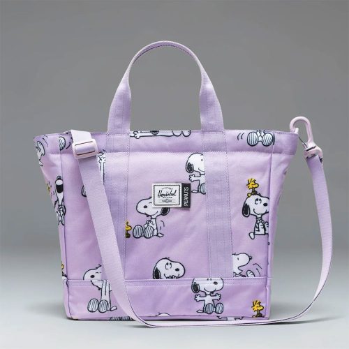 Lavender Herschel tote bag featuring a repeating pattern of Snoopy and Woodstock illustrations, complete with top carrying handles, an adjustable shoulder strap, and a Herschel x Peanuts woven brand label on the front pocket.